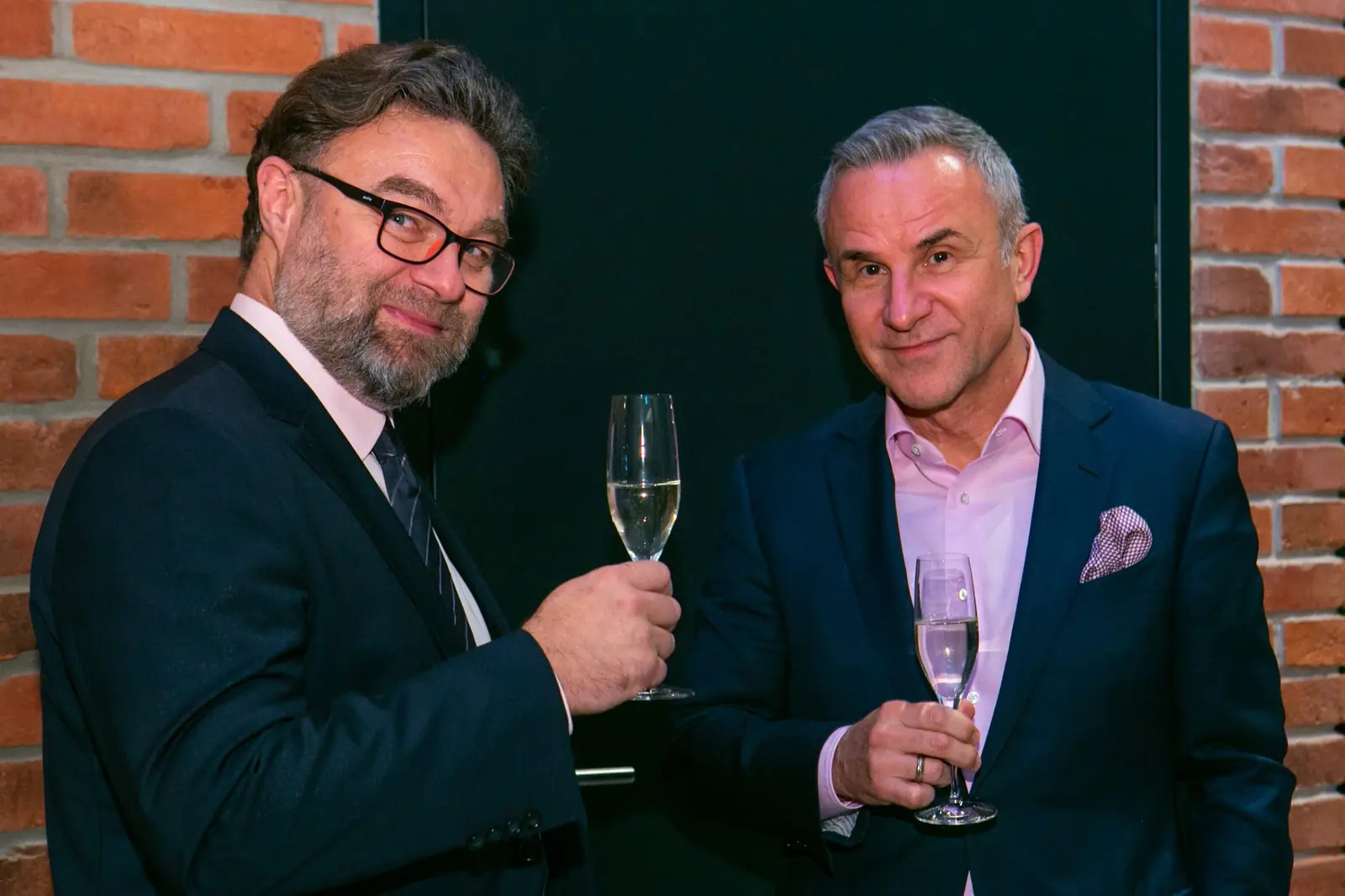 Two men in suits with glasses of champagne pose by a brick wall — DMR Gala X (2021)