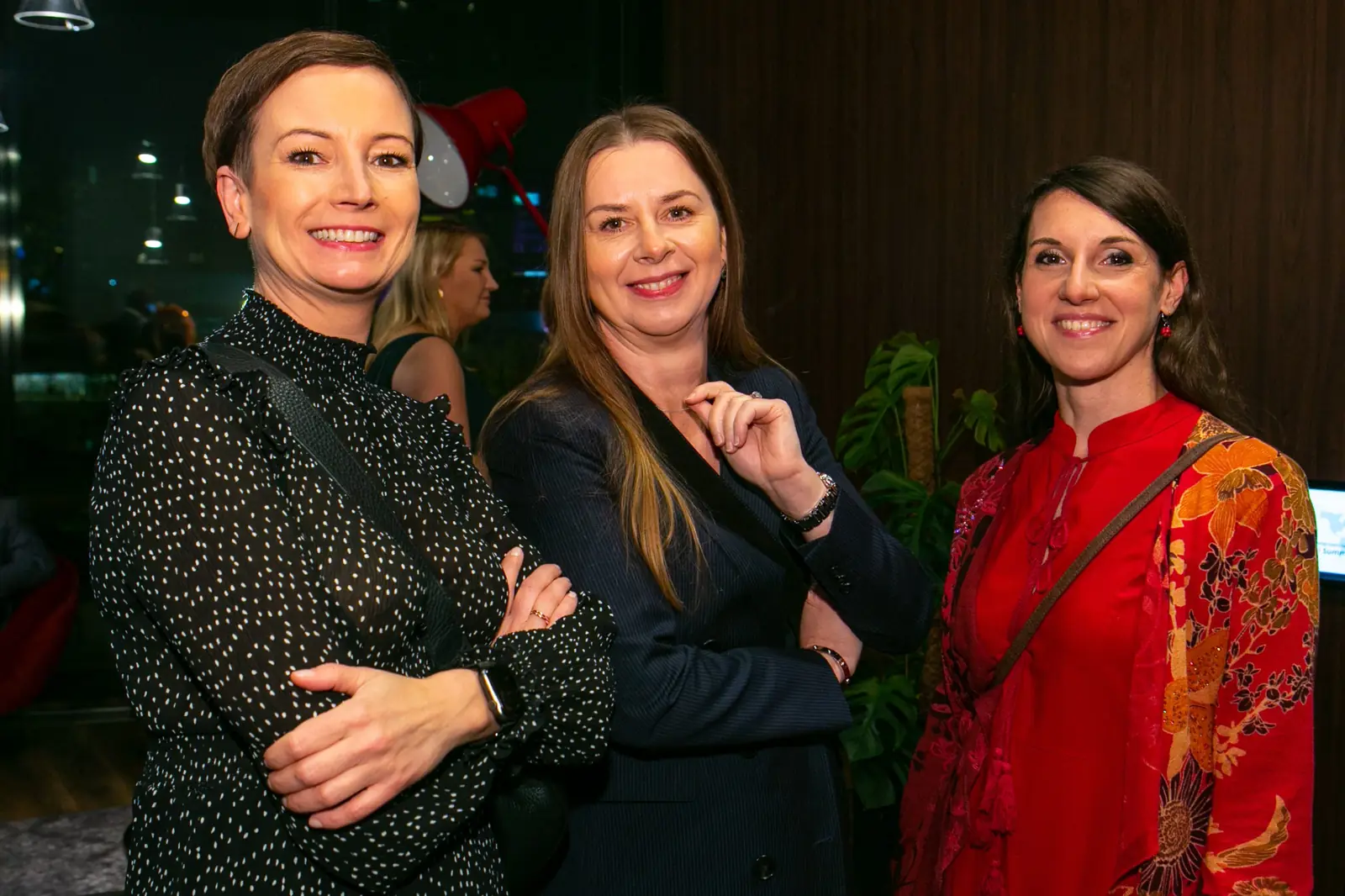 Three smiling women, one in a red dress with an oriental floral print, pose in the welcome area — DMR Gala X (2021)