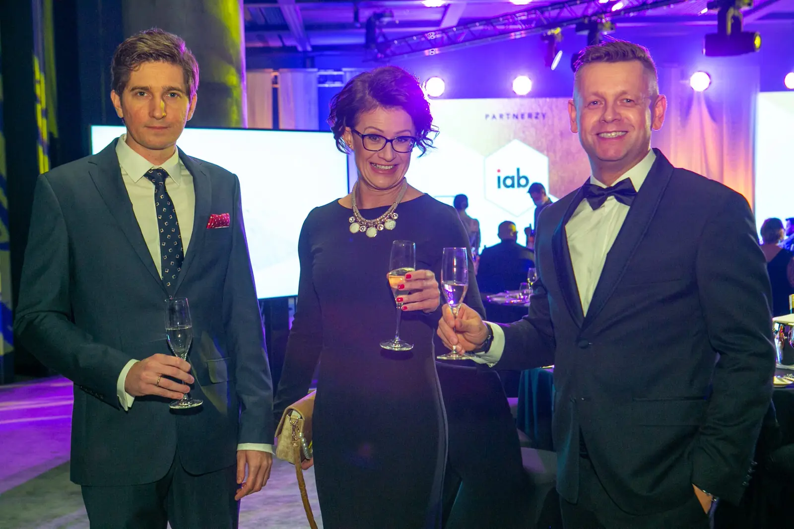 Host in a tuxedo with a glass of champagne poses with two guests by the partners' screen — DMR Gala X (2021)
