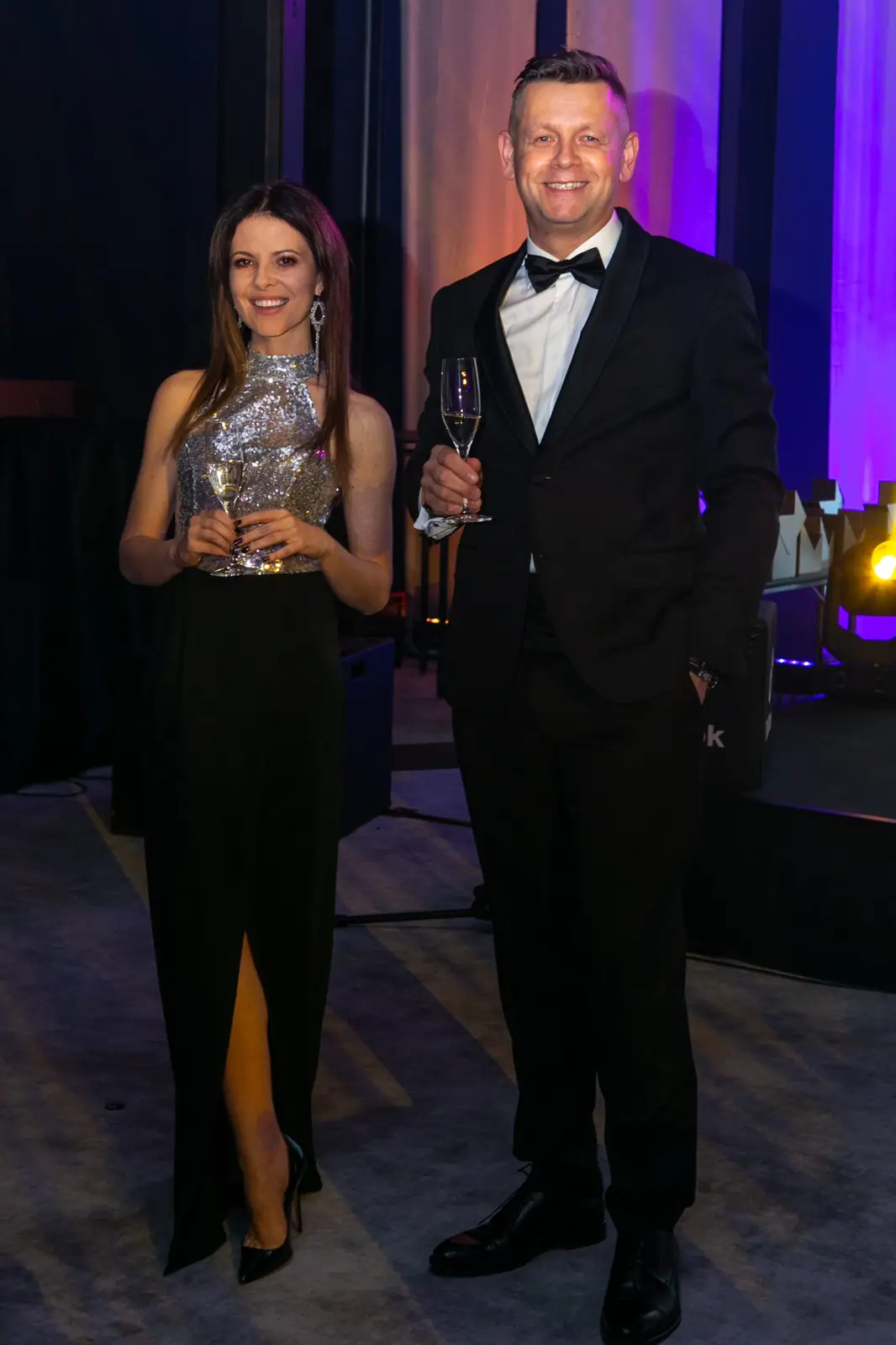Woman in a silver sequinned top and the host in a tuxedo pose with glasses of champagne — DMR Gala X (2021)