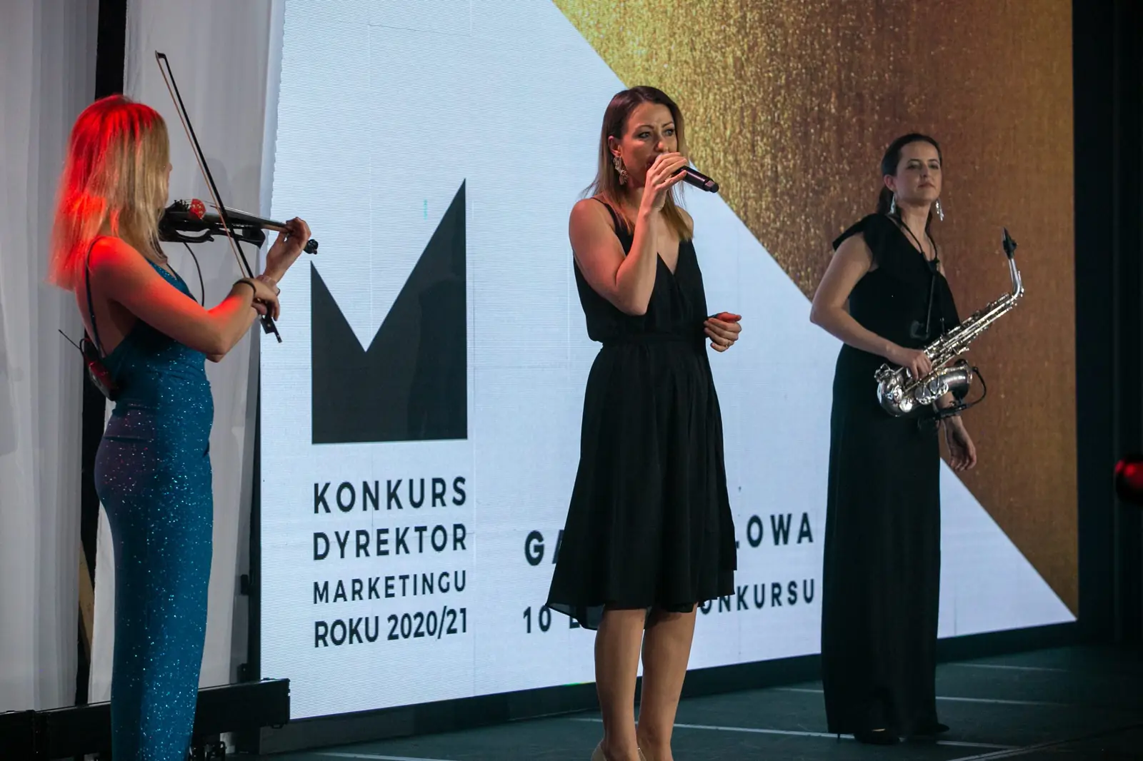 Singer in a black dress performs with a violinist and saxophonist beside the contest logo — DMR Gala X (2021)