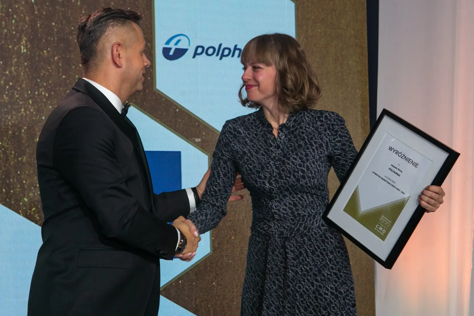 Host in a tuxedo congratulates a winner in a patterned dress who is holding her diploma — DMR Gala X (2021)