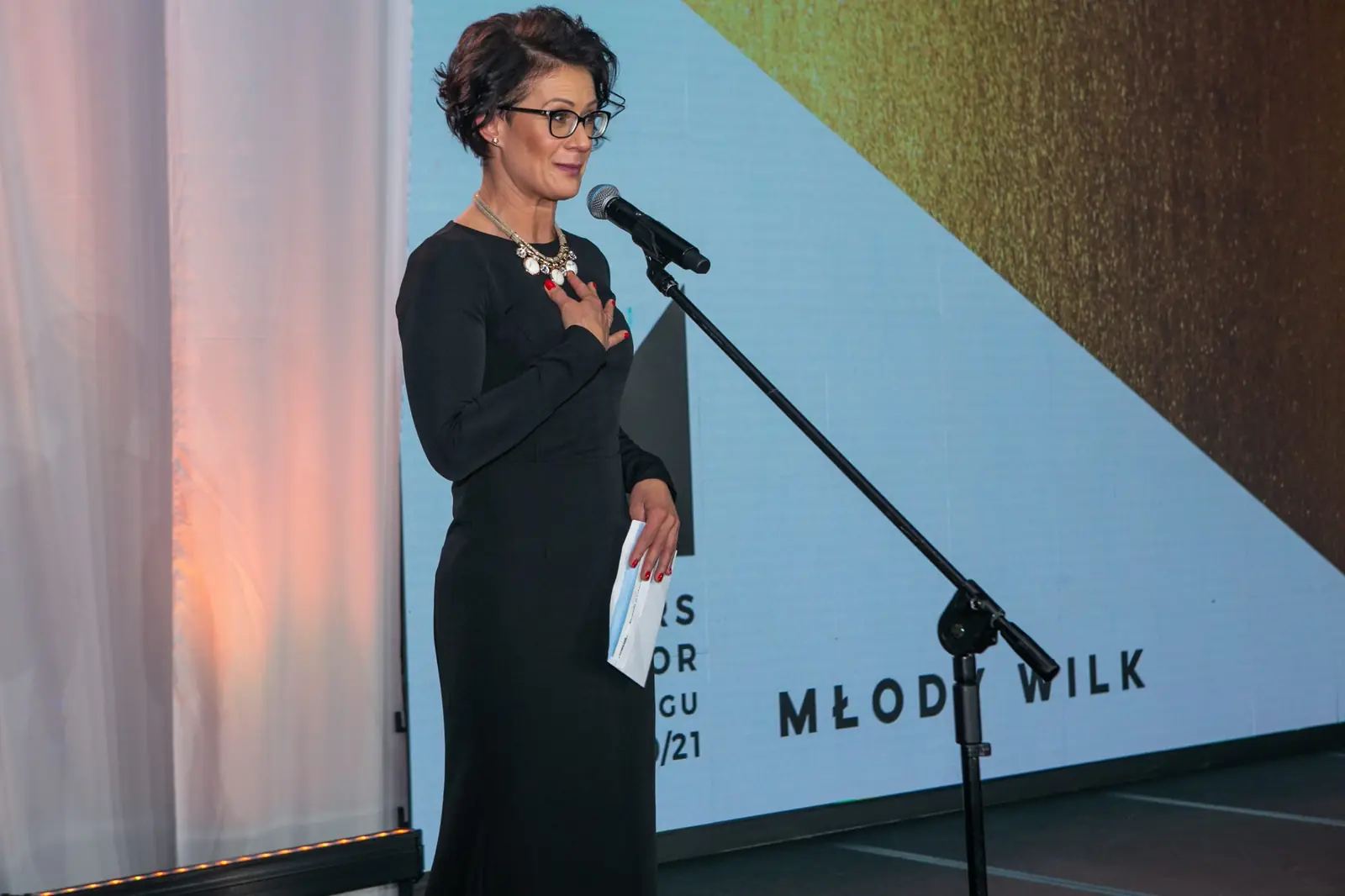 Presenter in a black dress, hand on heart, announces the Young Wolf award at the microphone — DMR Gala X (2021)