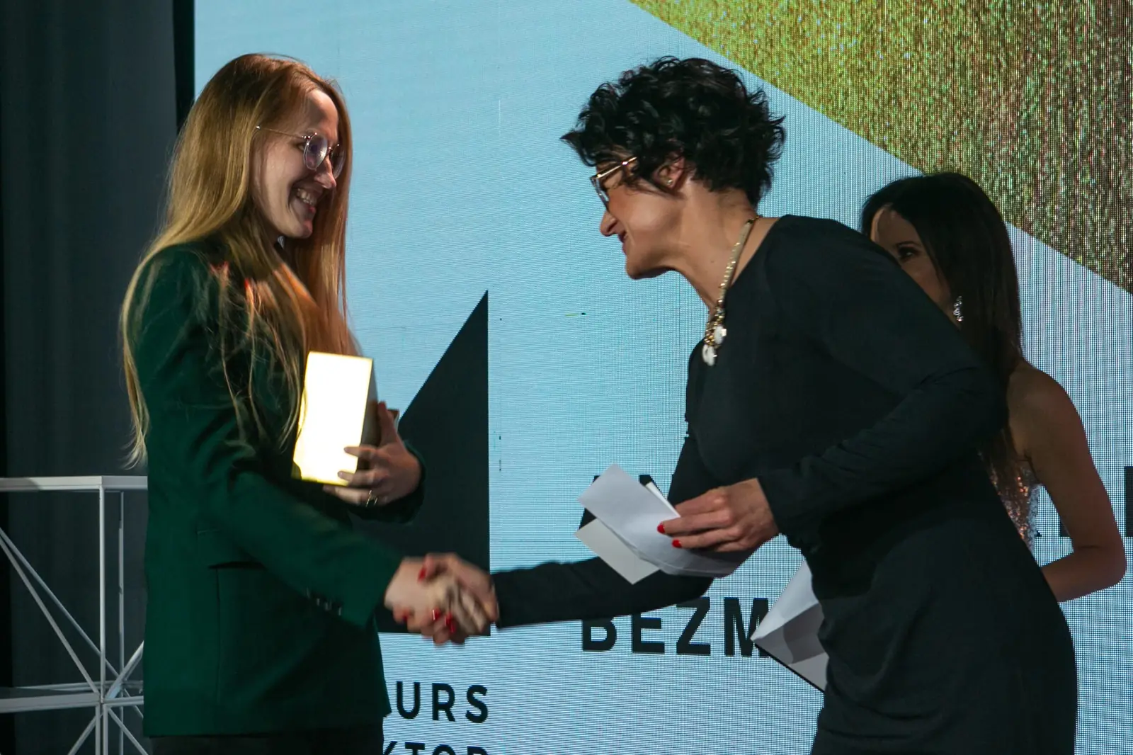 Laughing winner in green holding a statuette shakes hands with the presenter in glasses — DMR Gala X (2021)