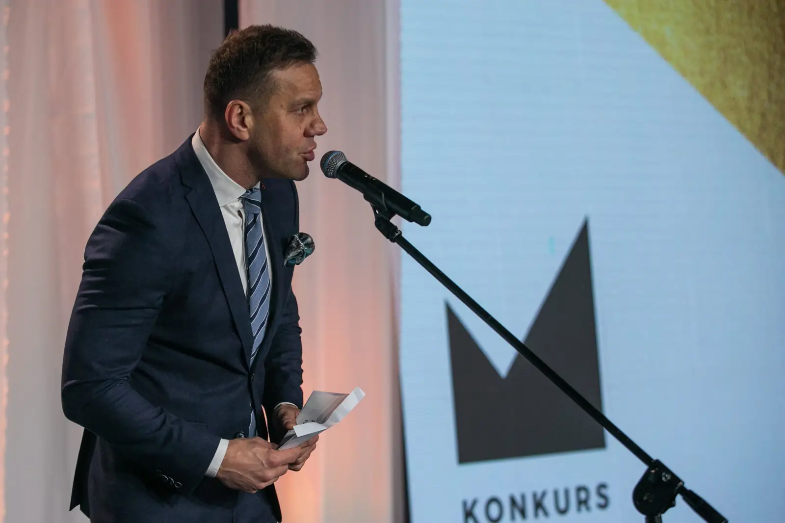 Man in a navy suit announces the next category into a stand microphone — DMR Gala X (2021)