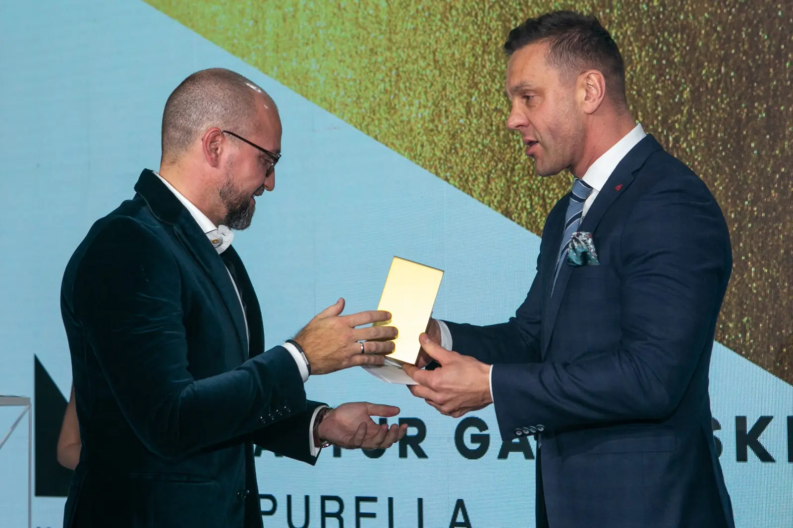 Presenter in a navy suit hands a golden statuette to a winner in a velvet jacket — DMR Gala X (2021)