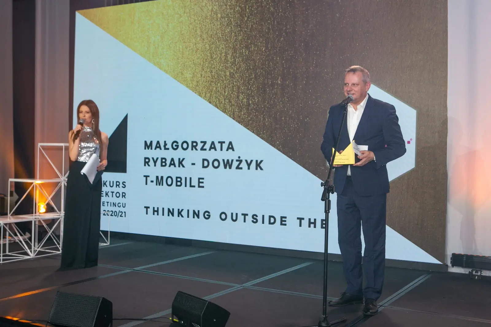 Wide stage shot: the co-host in a sparkling gown and a winner with a statuette at the microphone — DMR Gala X (2021)
