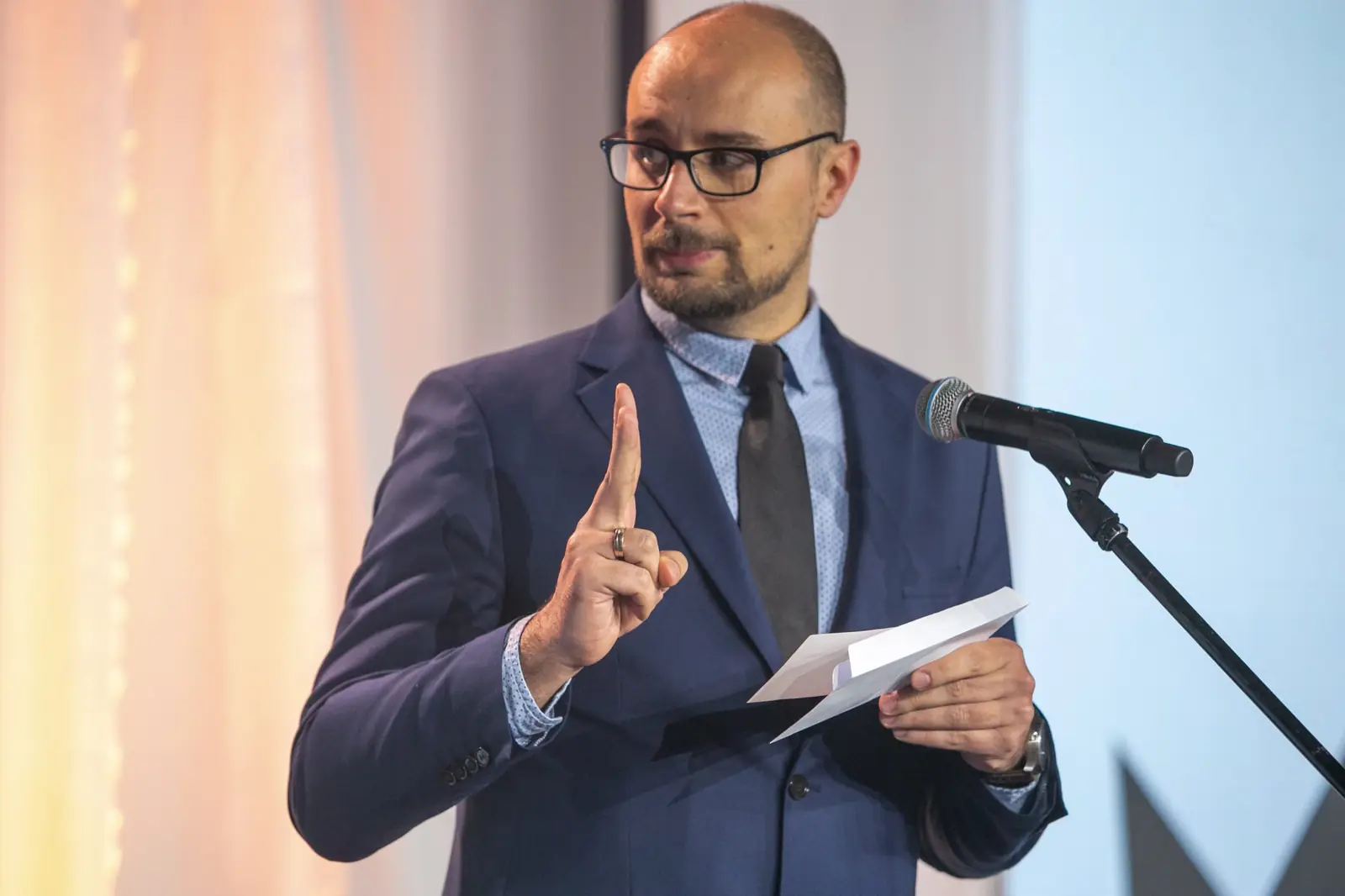 Mężczyzna w okularach unosi palec wskazujący, mówiąc do mikrofonu z kartką w dłoni — Gala DMR X (2021)