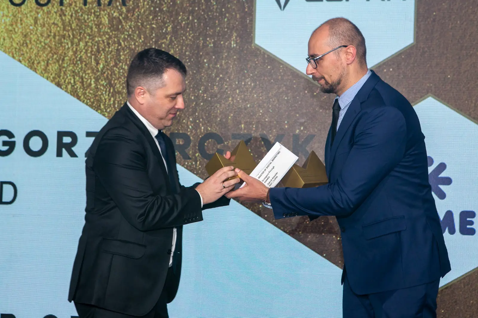 Laureat w czarnym garniturze sięga po statuetkę i kartę gratulacyjną od wręczającego w okularach — Gala DMR X (2021)