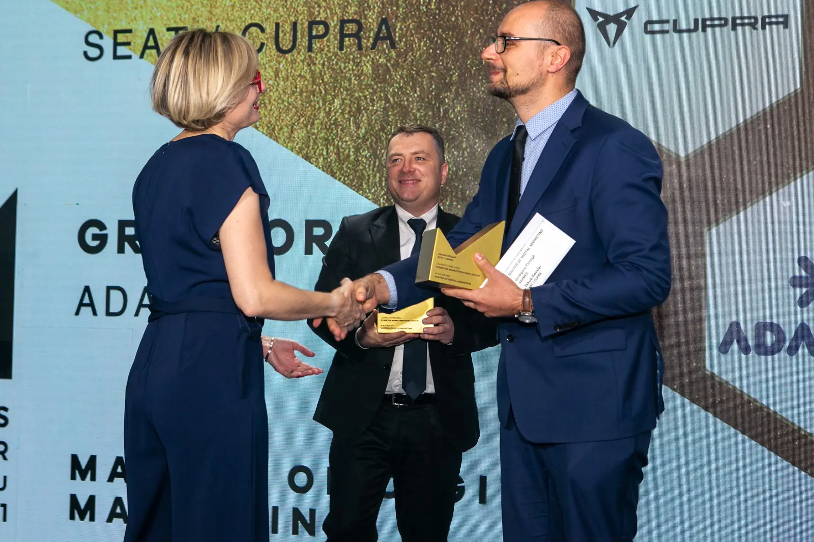 Woman in a navy jumpsuit shakes hands with the presenter holding a statuette, a winner behind — DMR Gala X (2021)