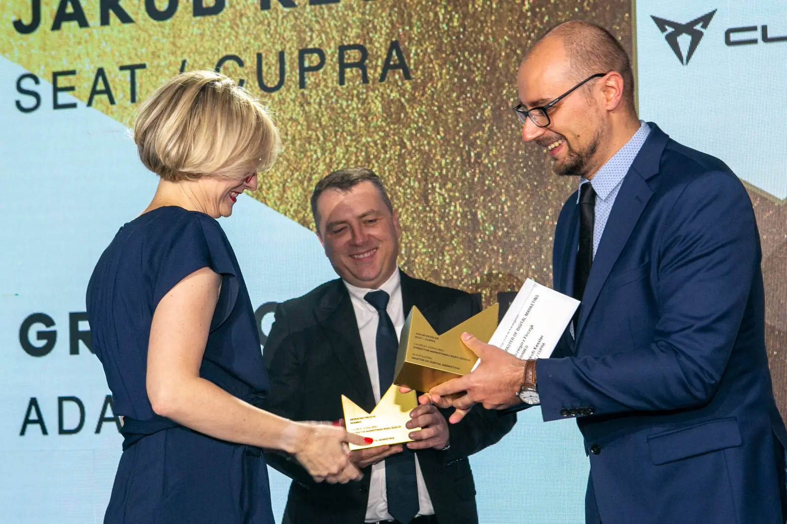 Woman in a navy dress receives a statuette from the presenter in glasses, another winner beside — DMR Gala X (2021)