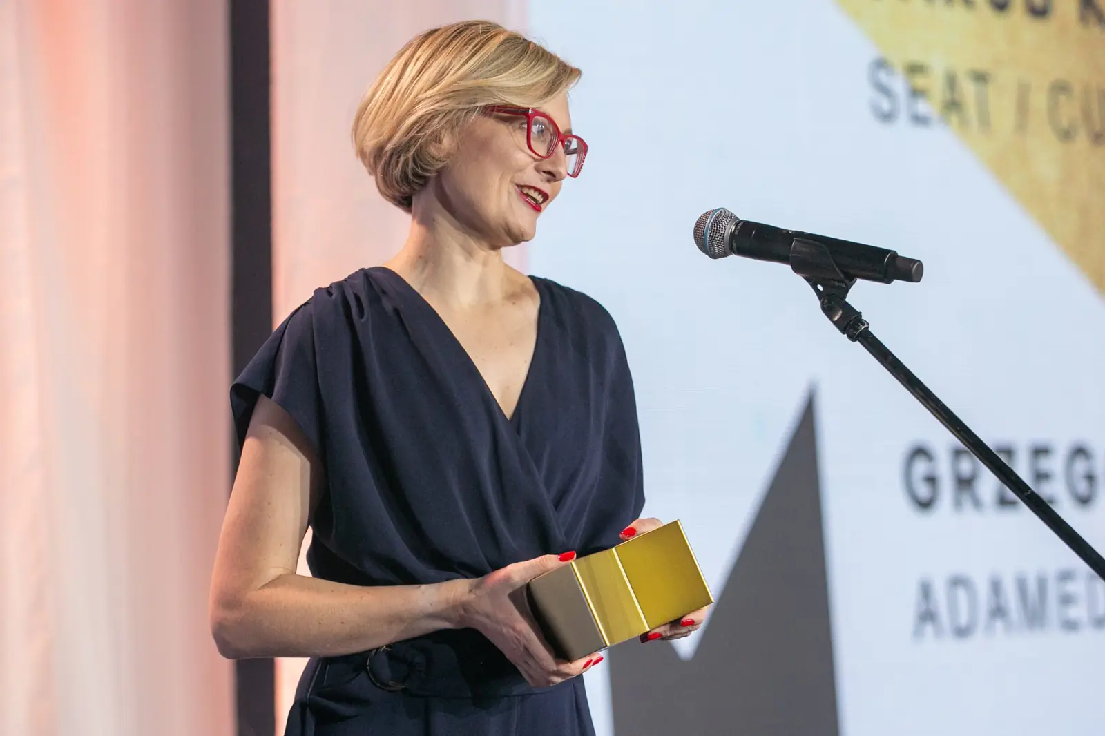 Woman in red glasses and a navy dress holds a golden statuette by the microphone — DMR Gala X (2021)