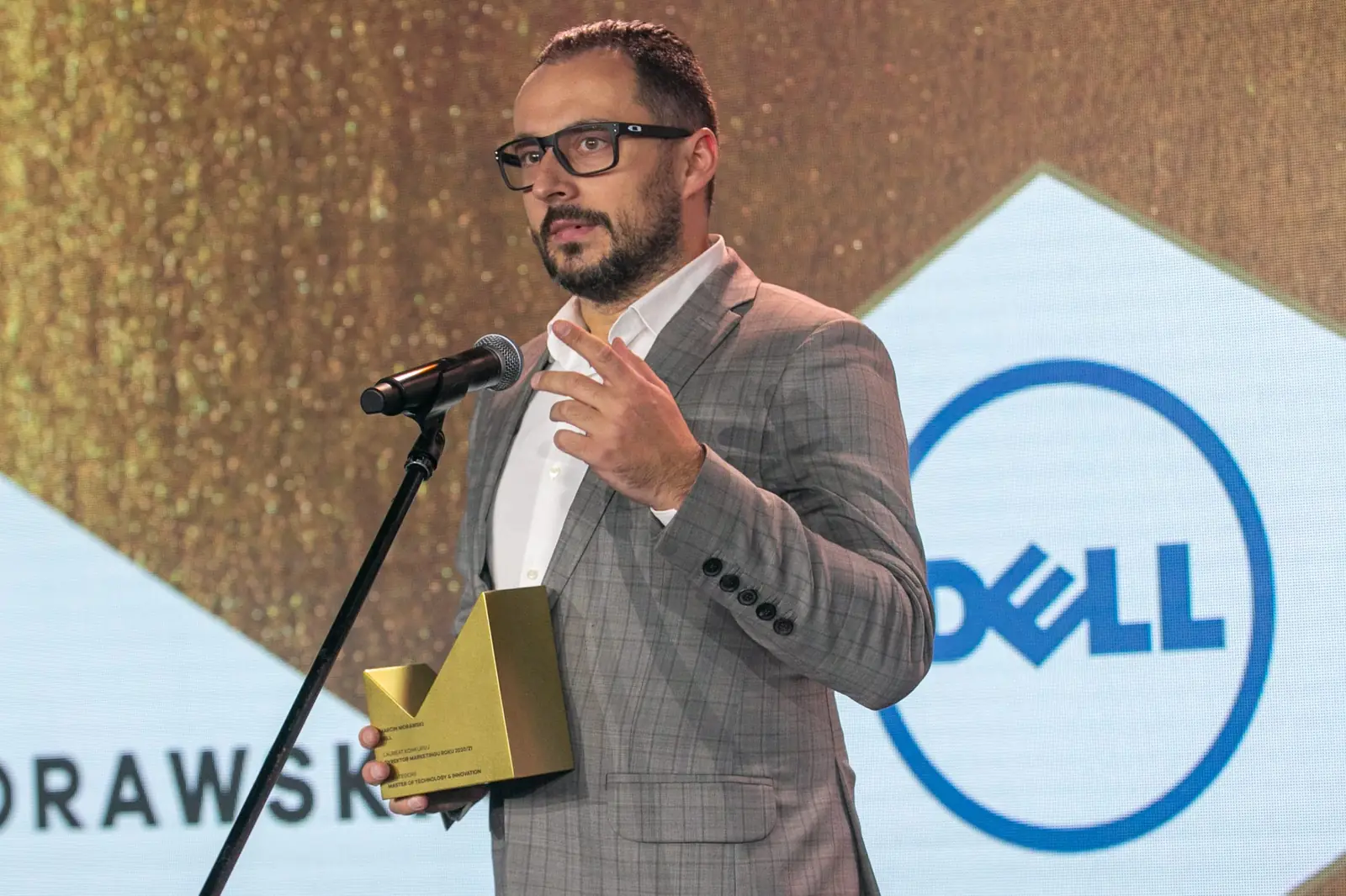 Laureat w okularach i szarej marynarce gestykuluje przy mikrofonie, trzymając statuetkę — Gala DMR X (2021)
