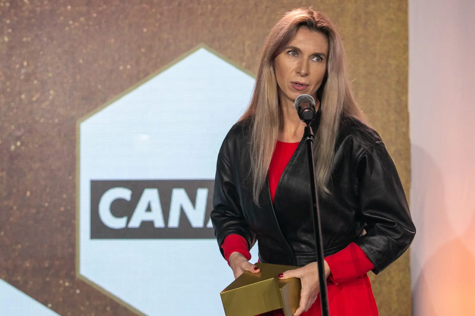 Winner in a leather jacket and red dress speaks at the microphone holding her statuette — DMR Gala X (2021)