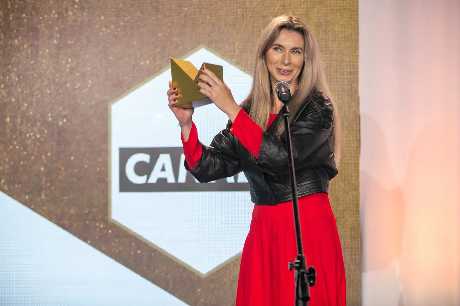 Winner in a red dress smiles as she lifts her golden statuette up by the microphone — DMR Gala X (2021)
