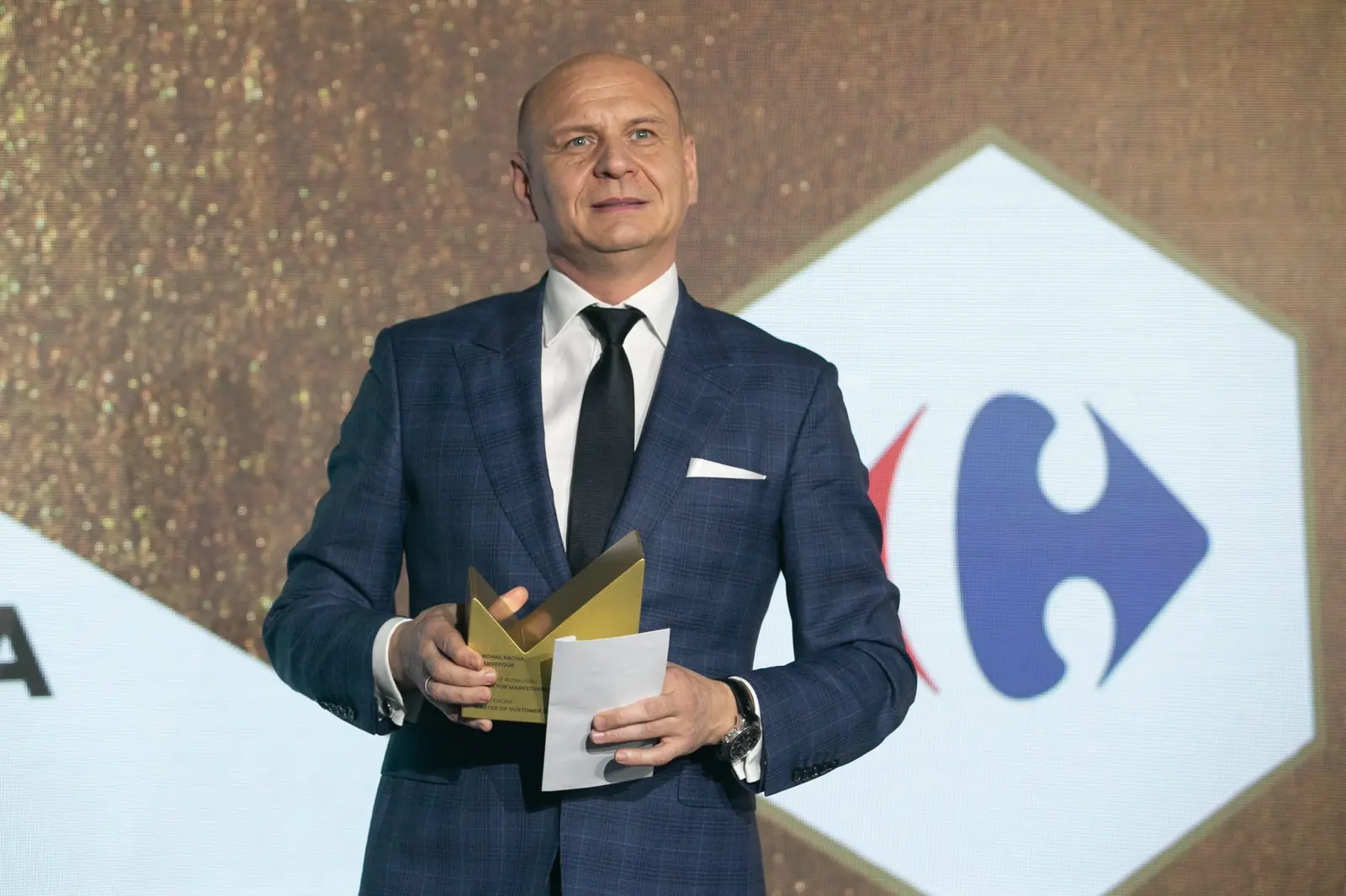 Smiling man in a navy checked suit holds a statuette and a card — DMR Gala X (2021)