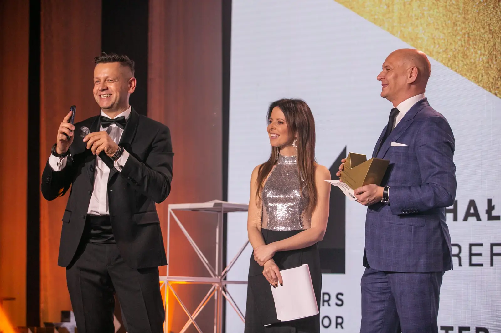 Prowadzący w smokingu i prowadząca w srebrnej bluzce na scenie z mężczyzną ze statuetką — Gala DMR X (2021)