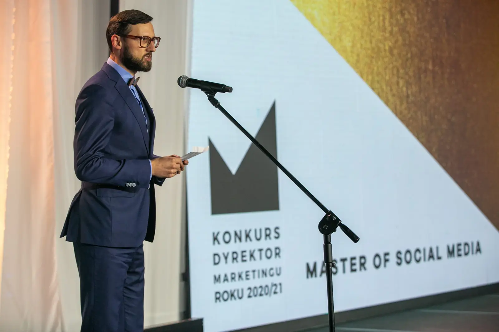 Wręczający w granatowym garniturze zapowiada przy mikrofonie kategorię Master of Social Media — Gala DMR X (2021)