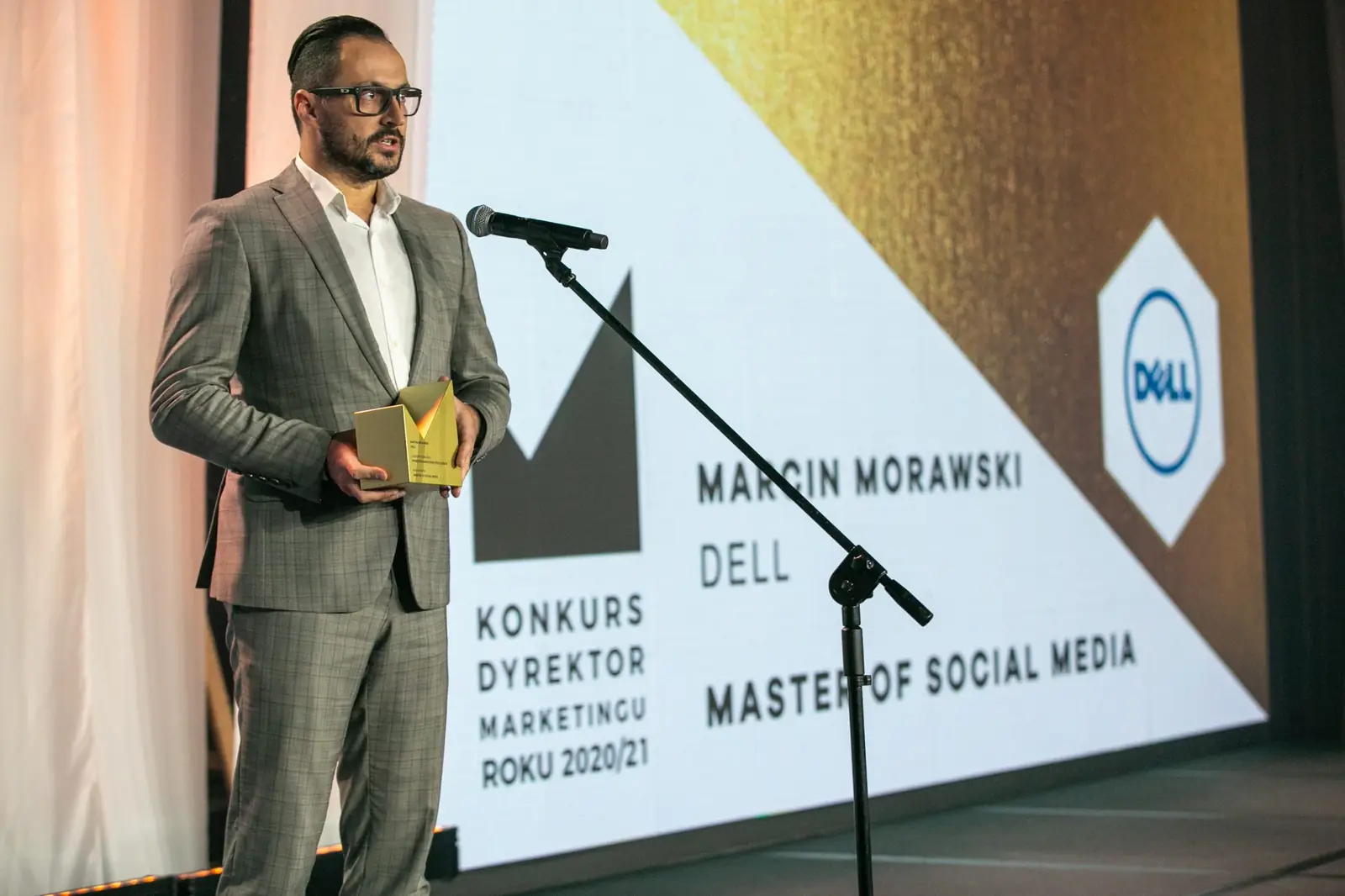 Laureat w szarym garniturze w kratę przemawia ze statuetką w kategorii Master of Social Media — Gala DMR X (2021)