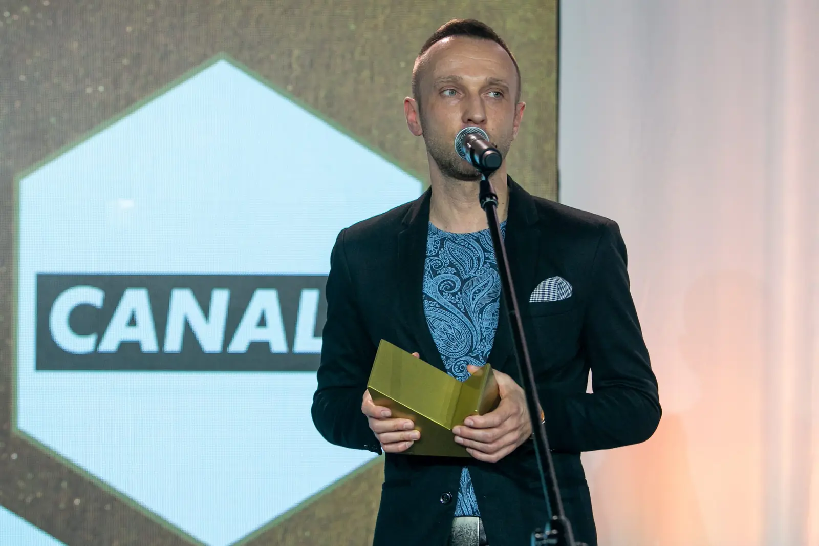 Laureat w ciemnej marynarce i wzorzystej koszuli mówi do mikrofonu ze statuetką w dłoniach — Gala DMR X (2021)