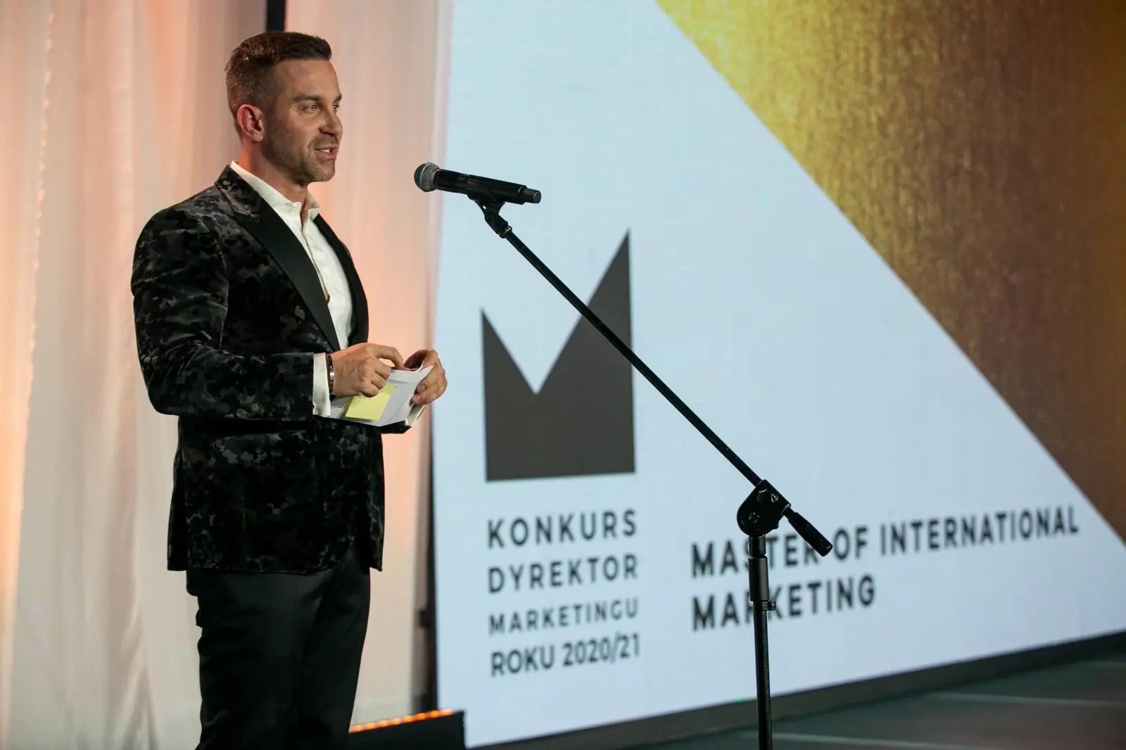 Mężczyzna w czarnej wzorzystej marynarce zapowiada kategorię Master of International Marketing — Gala DMR X (2021)