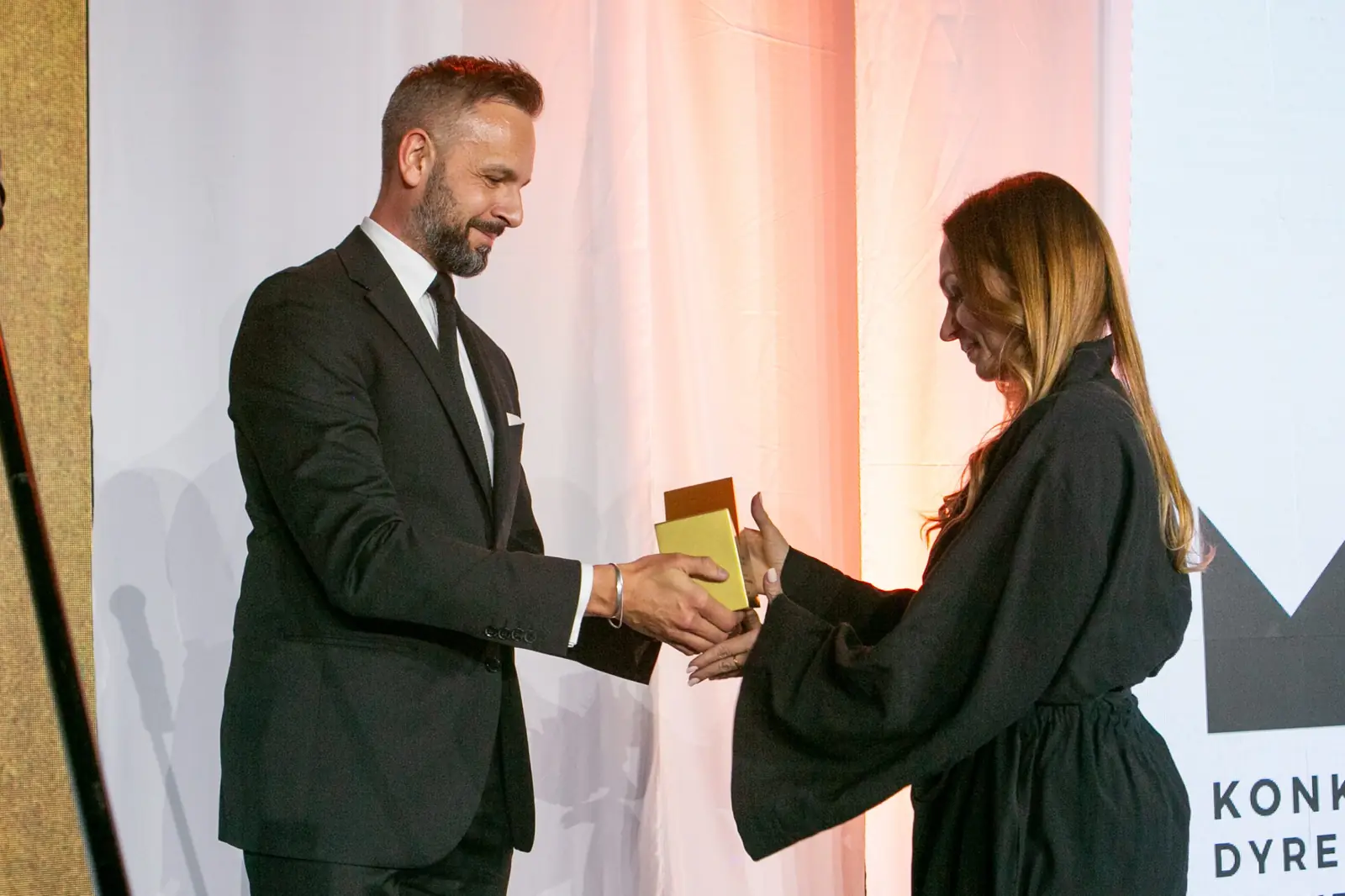 Bearded presenter hands the statuette to a winner in a black dress with wide sleeves — DMR Gala X (2021)