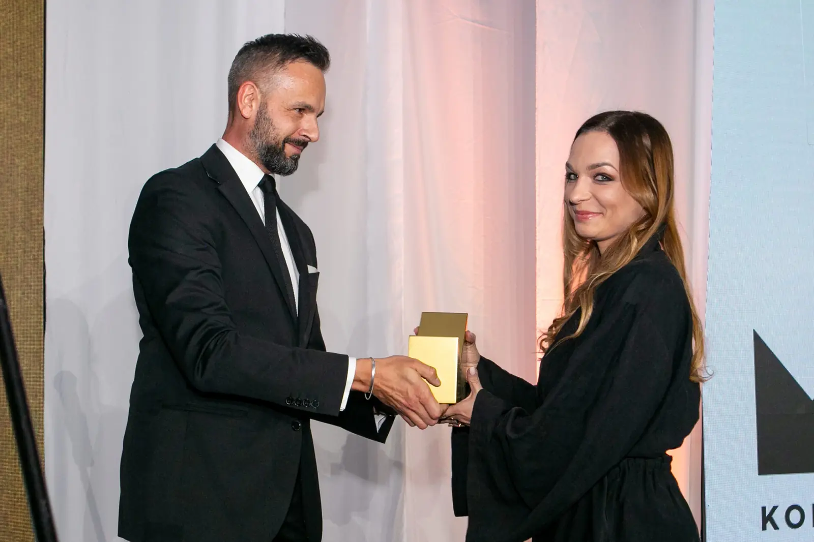 Laureatka o długich włosach z uśmiechem przyjmuje statuetkę od brodatego wręczającego — Gala DMR X (2021)