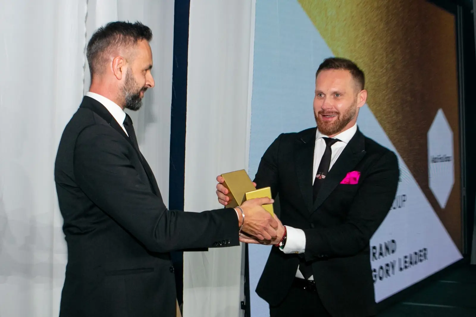 Winner with a pink pocket square accepts the statuette from the presenter in a black suit — DMR Gala X (2021)
