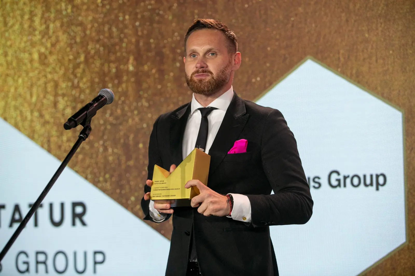 Bearded winner in a black suit poses with a golden statuette in front of a company logo — DMR Gala X (2021)