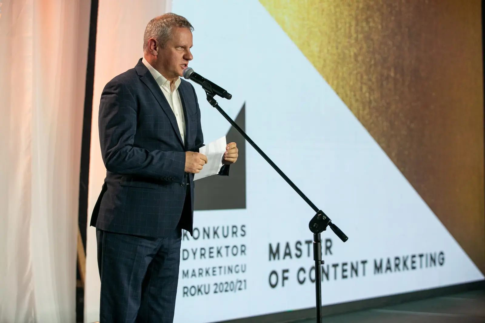 Szpakowaty wręczający zapowiada przy mikrofonie kategorię Master of Content Marketing — Gala DMR X (2021)