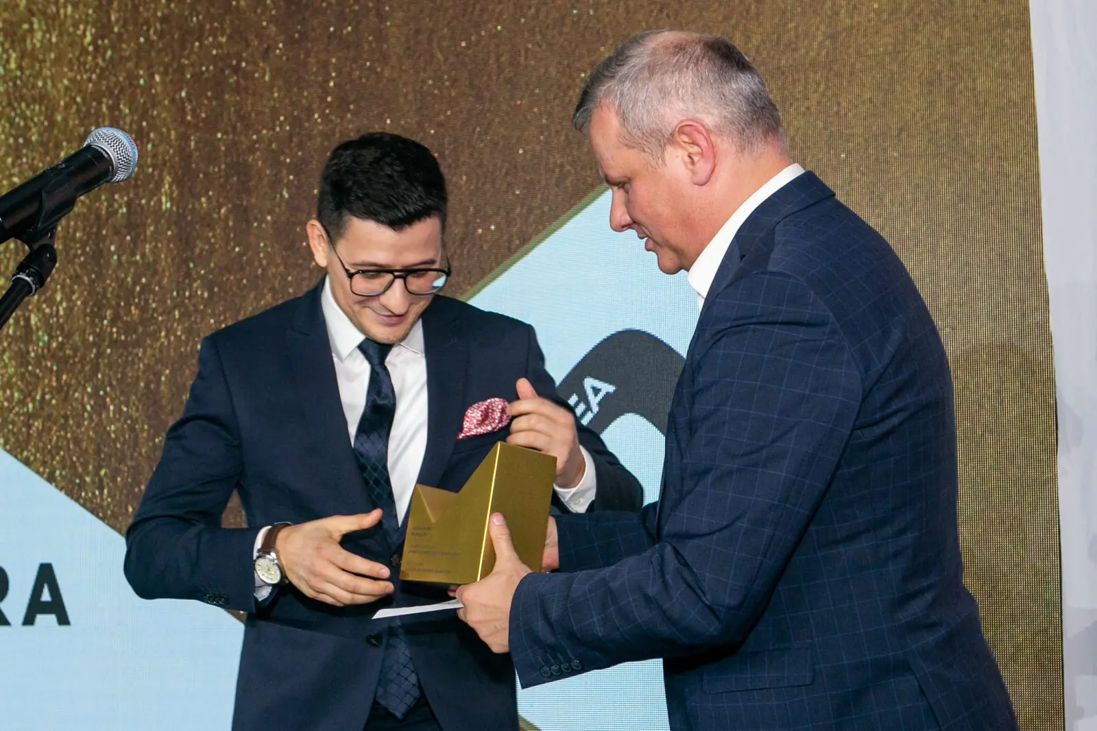 Młody laureat w okularach odbiera złotą statuetkę od szpakowatego wręczającego — Gala DMR X (2021)