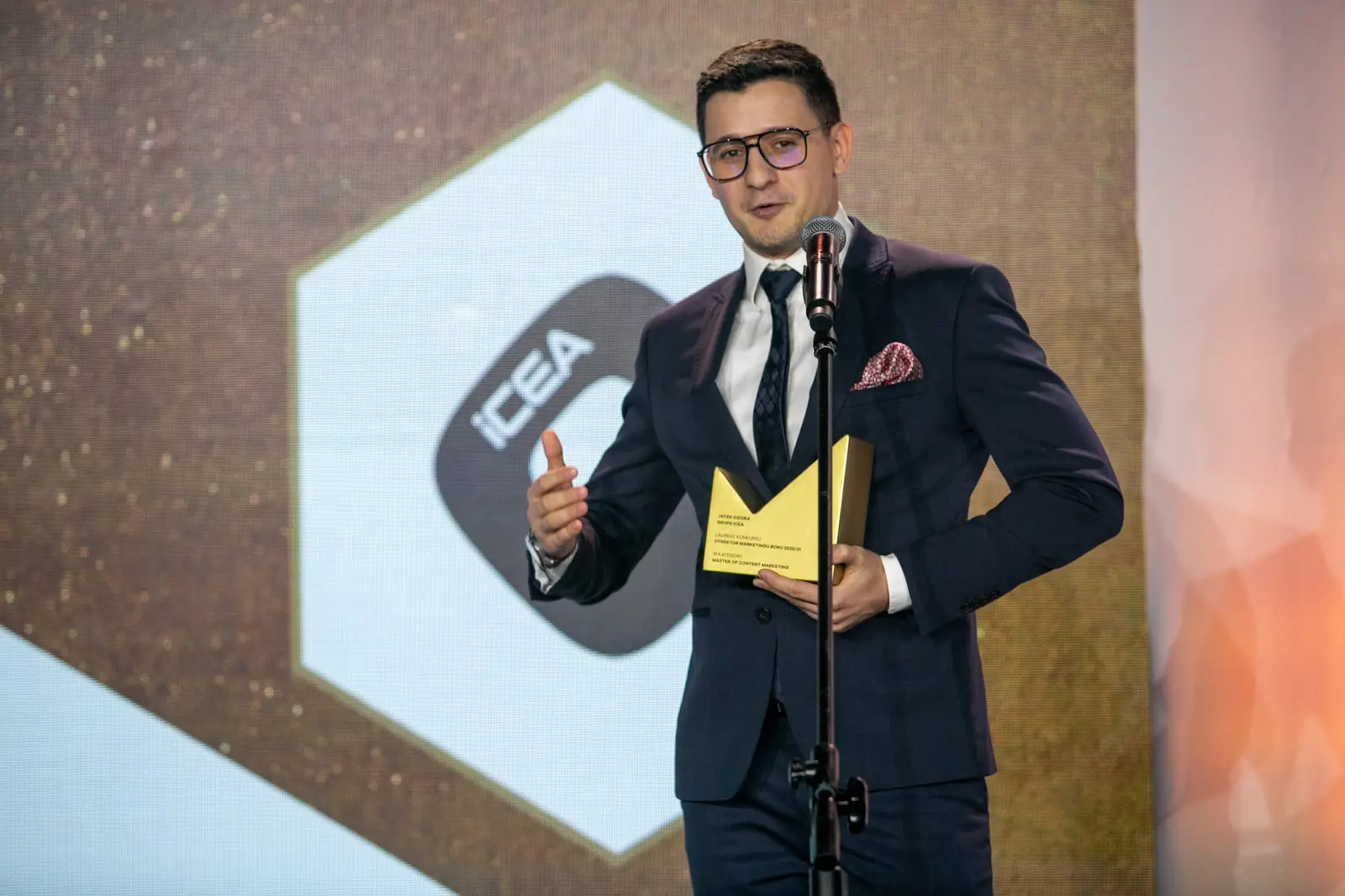 Laureat w okularach i granatowym garniturze przemawia, trzymając statuetkę przy mikrofonie — Gala DMR X (2021)