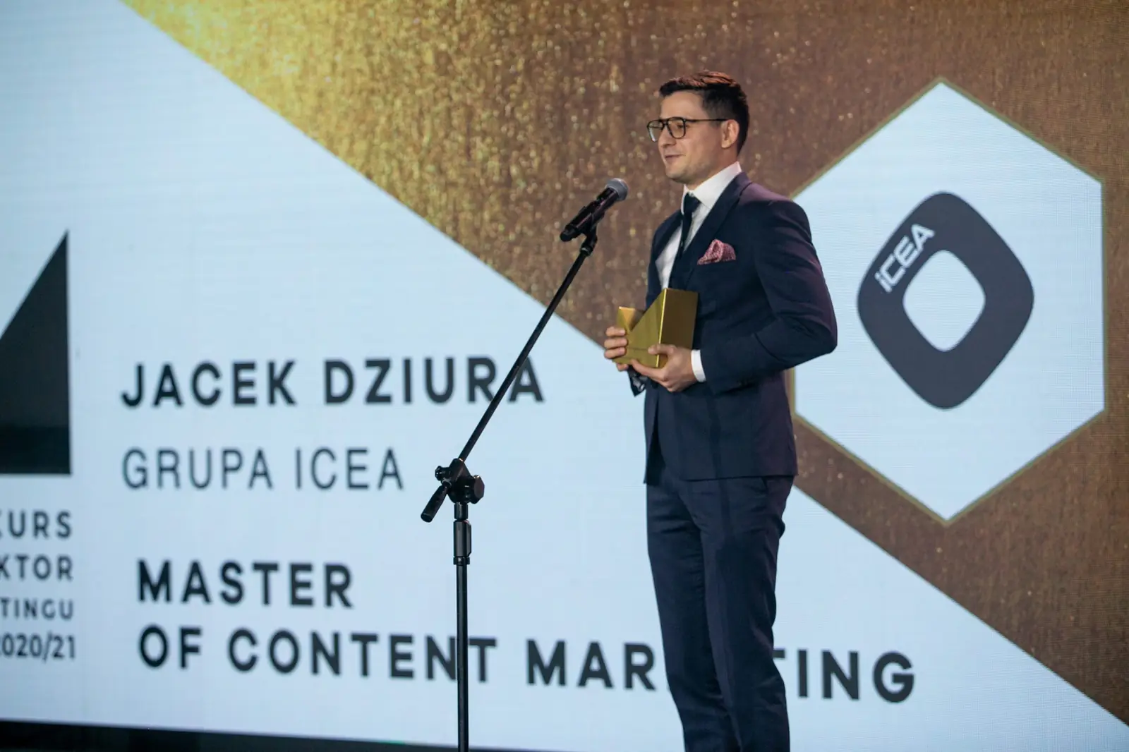 Laureat w kategorii Master of Content Marketing stoi przy mikrofonie obok ekranu z nazwą nagrody — Gala DMR X (2021)