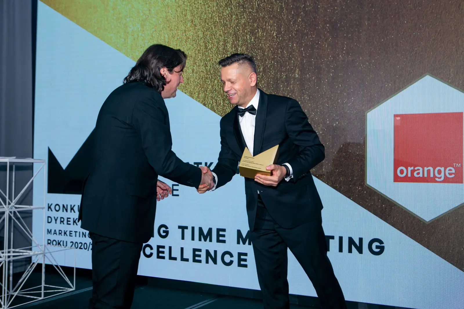 Host in a tuxedo presents the long-time excellence award to a man with long hair — DMR Gala X (2021)