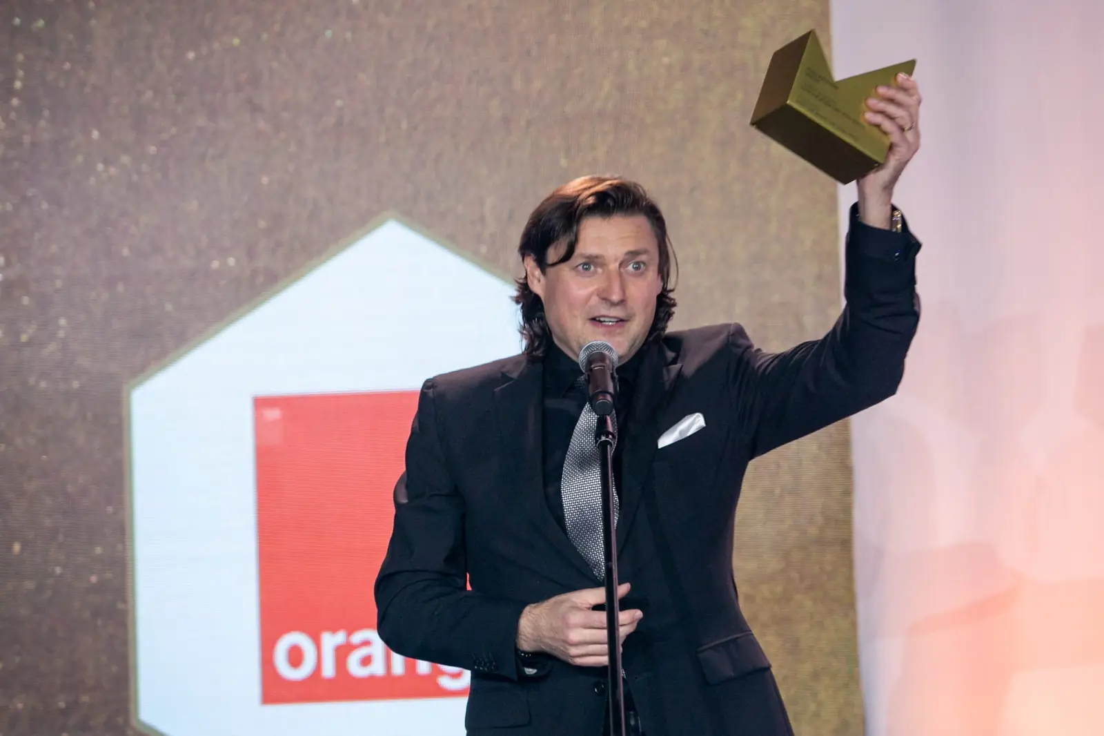 Laureat z długimi włosami unosi statuetkę wysoko nad głowę, mówiąc do mikrofonu — Gala DMR X (2021)