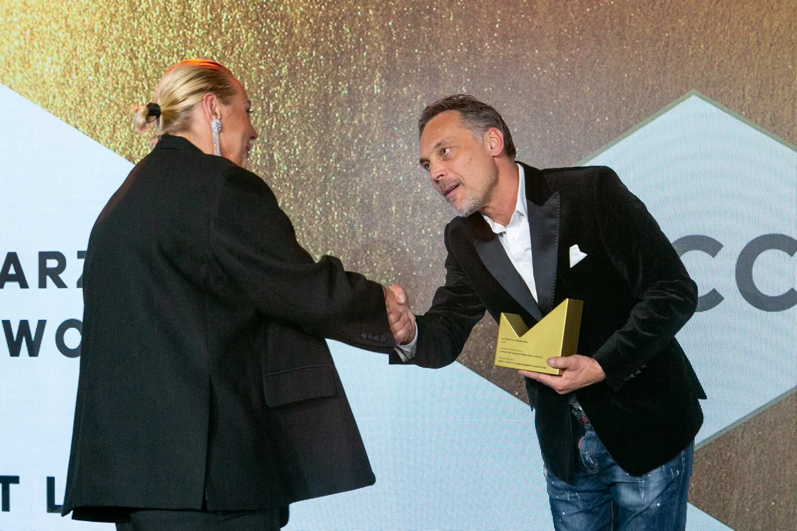 Winner with fair hair in a bun, wearing an oversized blazer, shakes the presenter's hand — DMR Gala X (2021)