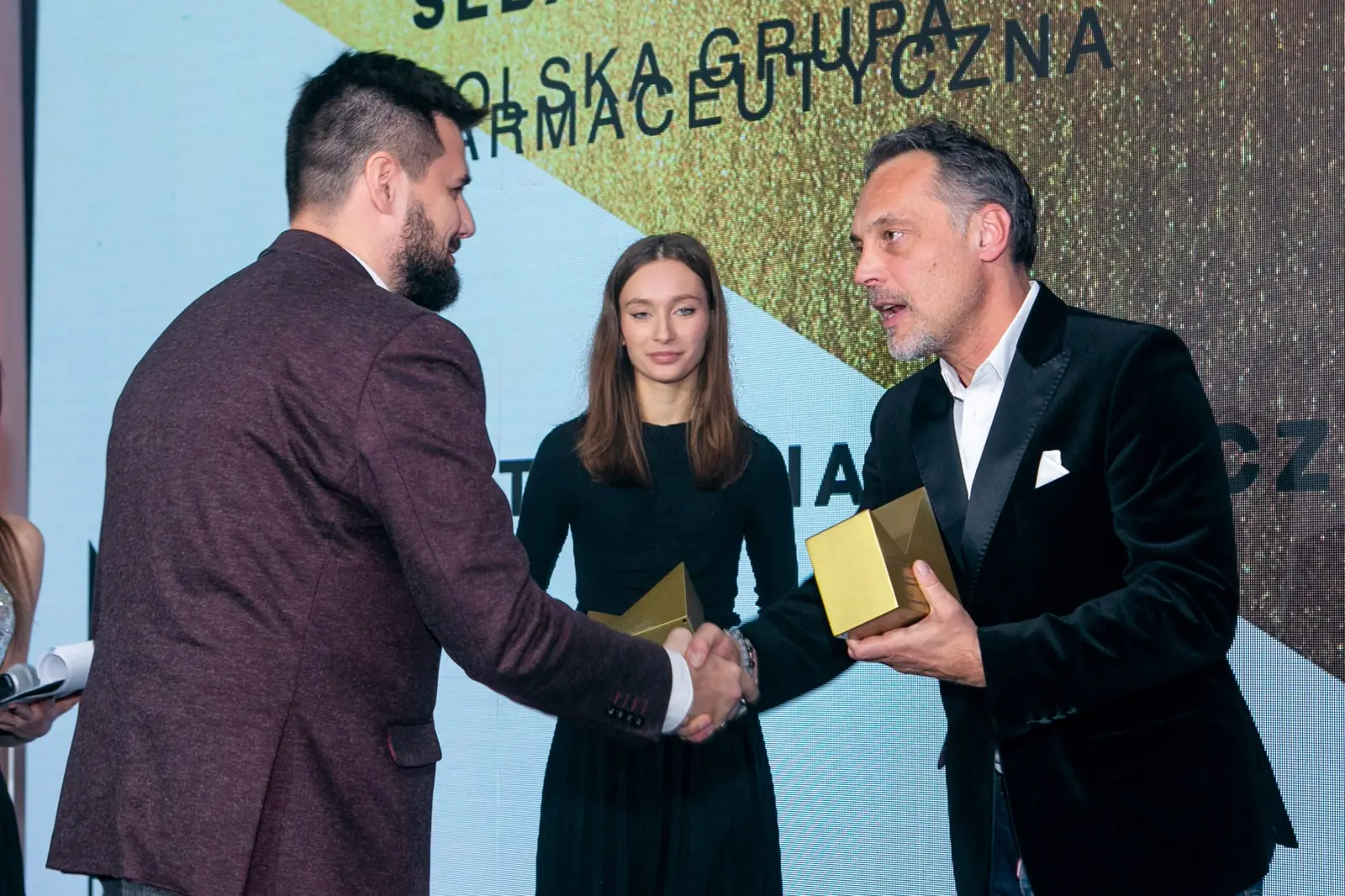 Brodaty laureat w bordowej marynarce ściska dłoń wręczającego, obok młoda kobieta ze statuetką — Gala DMR X (2021)