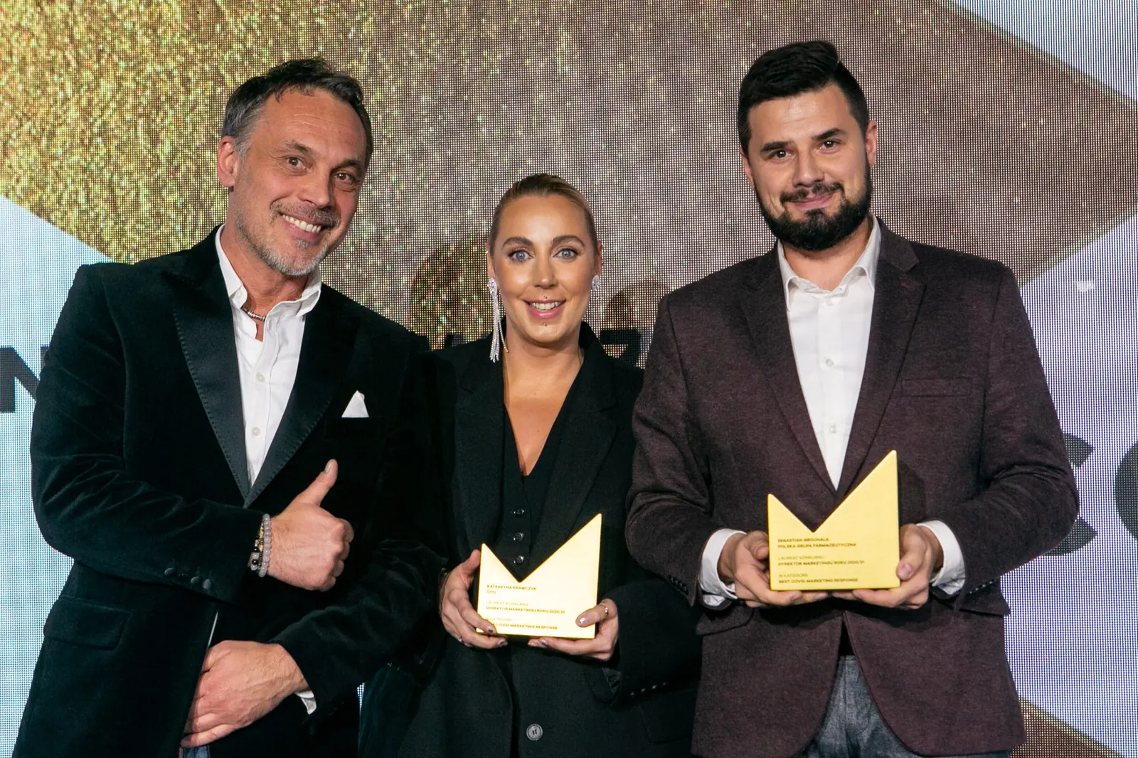 Wręczający pokazuje kciuk w górę obok dwojga laureatów ze złotymi statuetkami — Gala DMR X (2021)