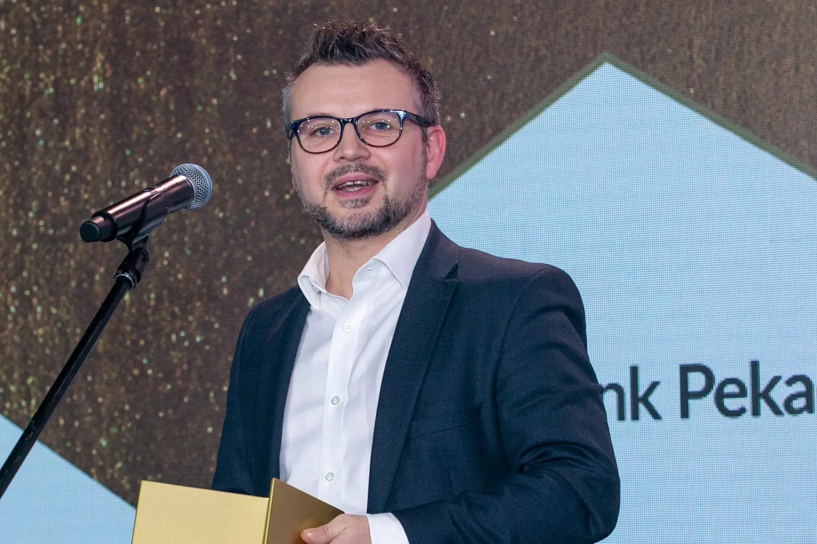 Uśmiechnięty laureat w okularach i ciemnej marynarce mówi do mikrofonu na tle logo firmy — Gala DMR X (2021)