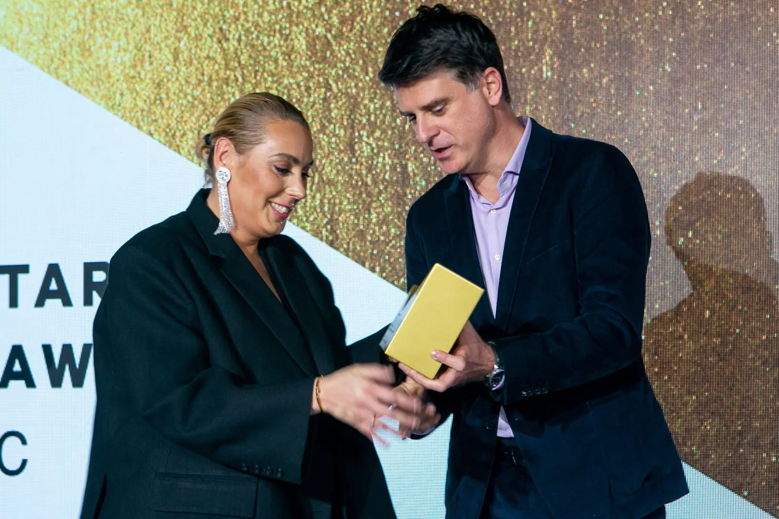Winner wearing large earrings and the presenter hold a golden statuette together — DMR Gala X (2021)