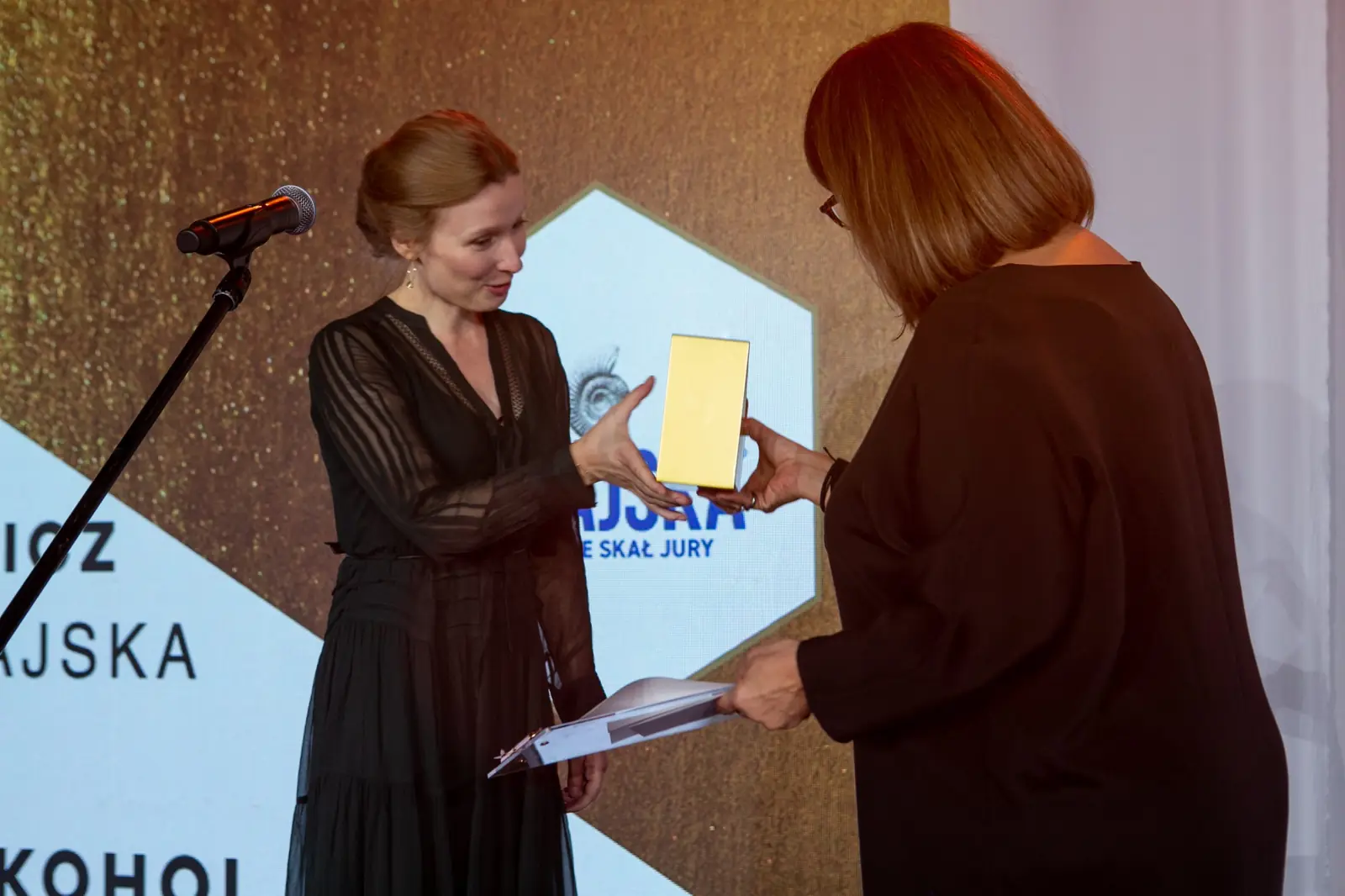 Laureatka w czarnej sukience z prześwitami odbiera statuetkę od wręczającej w okularach — Gala DMR X (2021)