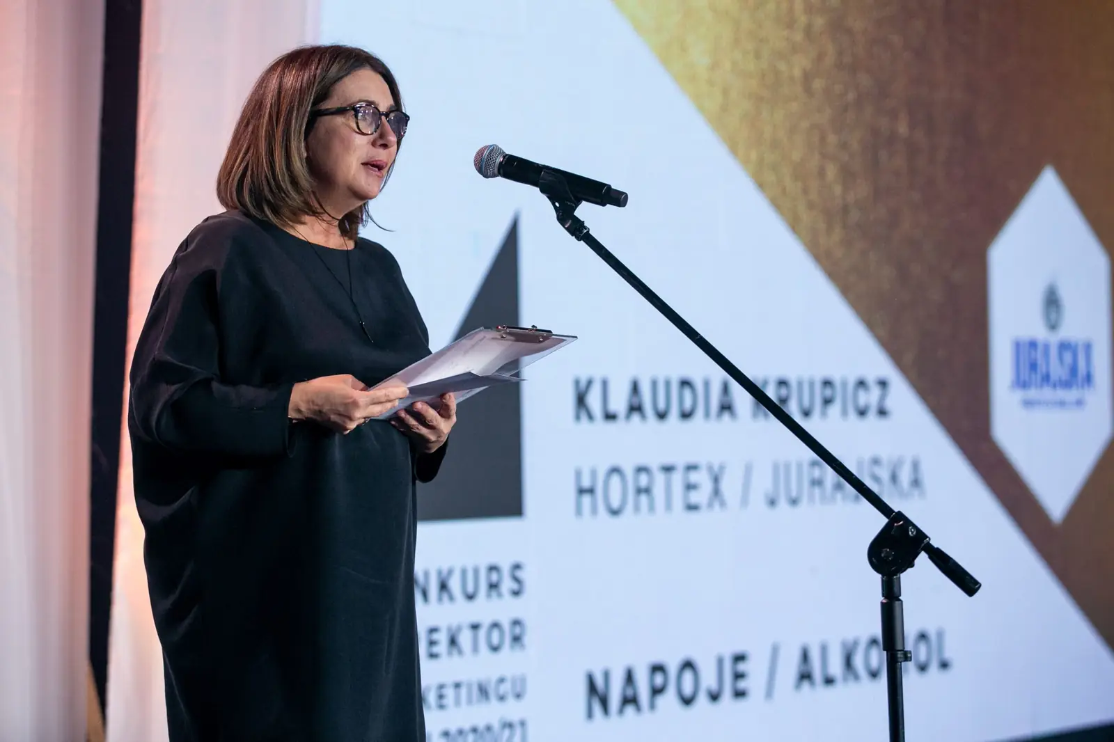 Wręczająca w czarnej tunice odczytuje werdykt przy mikrofonie obok nazwy kategorii — Gala DMR X (2021)