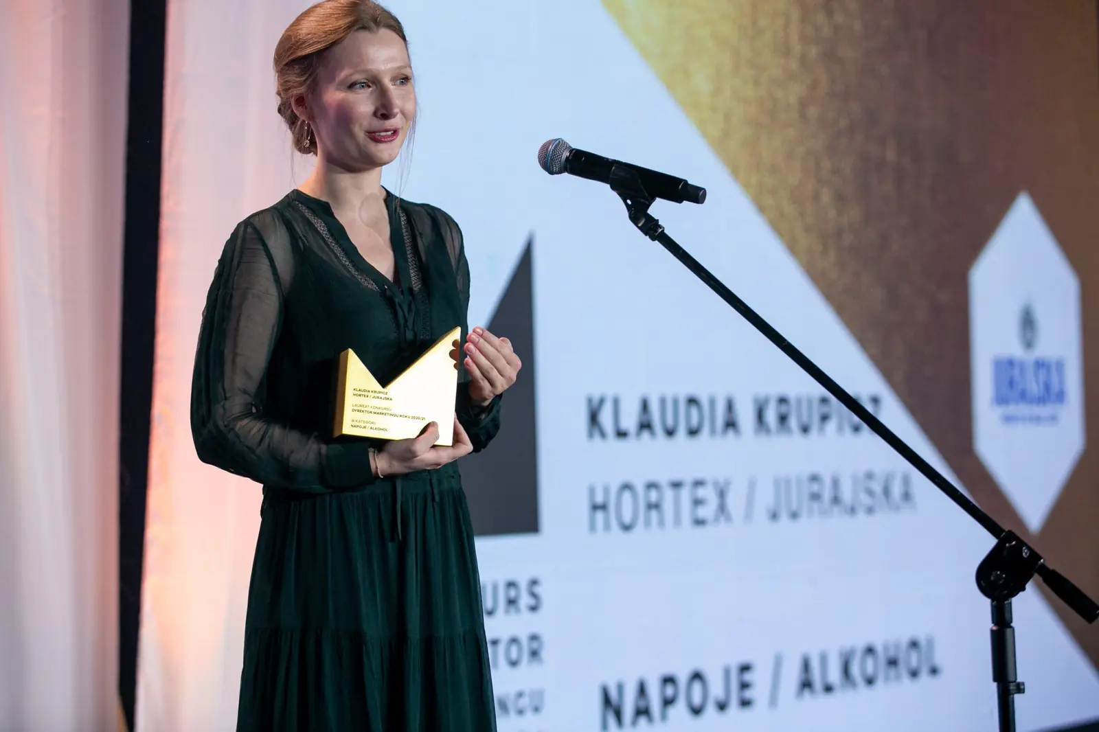 Laureatka z rudawym kokiem stoi ze statuetką obok ekranu z nazwą kategorii — Gala DMR X (2021)