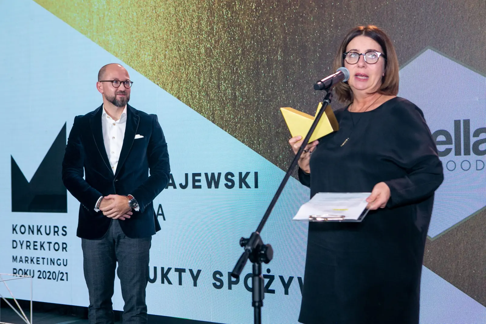 Wręczająca ze statuetką mówi do mikrofonu, a laureat w aksamitnej marynarce czeka obok — Gala DMR X (2021)
