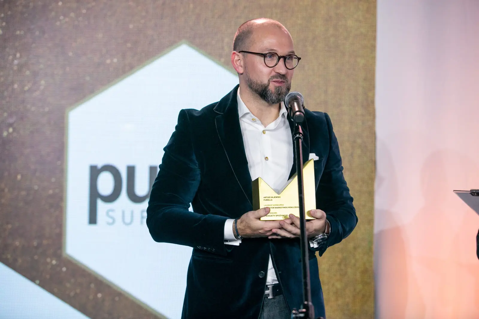 Brodaty laureat w okularach i aksamitnej marynarce dziękuje ze statuetką przy mikrofonie — Gala DMR X (2021)