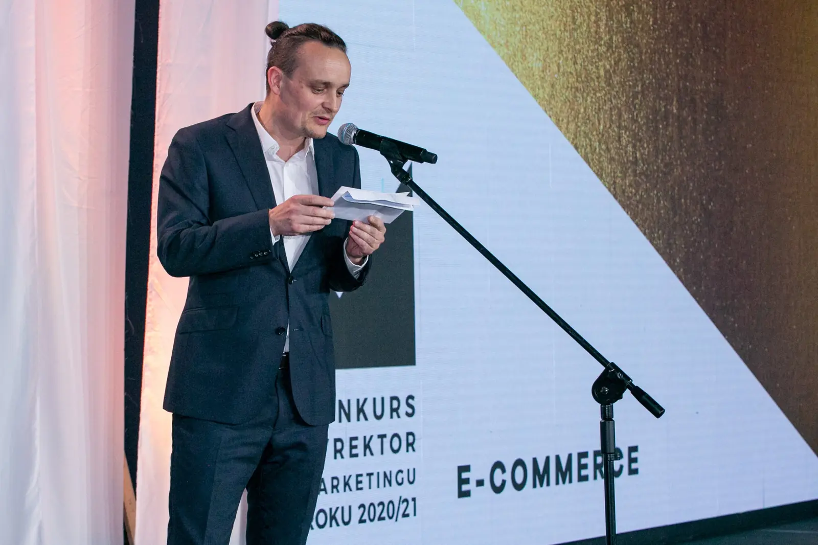 Wręczający z kokiem na czubku głowy zapowiada przy mikrofonie kategorię e-commerce — Gala DMR X (2021)