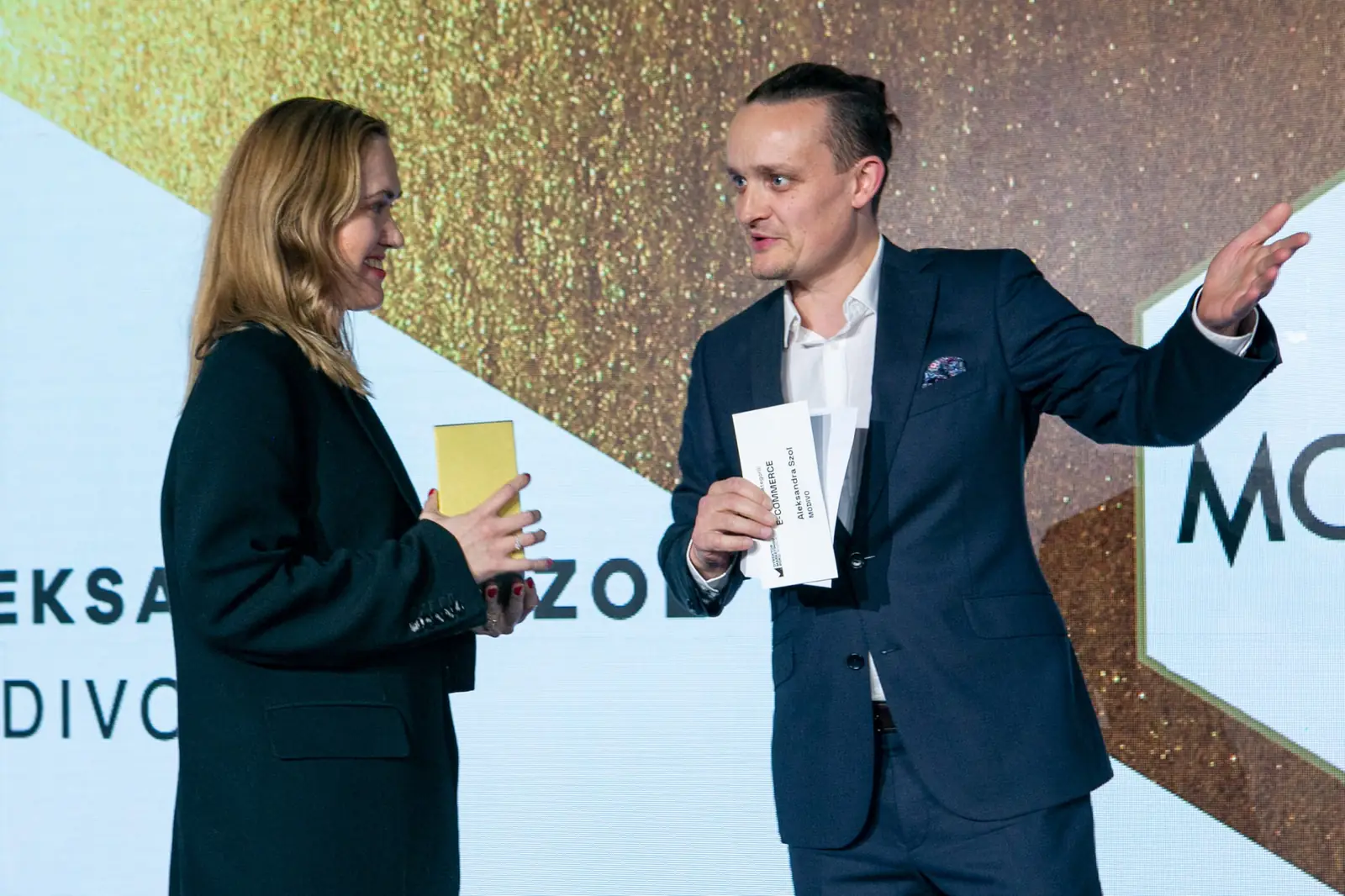 Wręczający wskazuje dłonią ekran, a laureatka ze statuetką stoi tuż obok — Gala DMR X (2021)