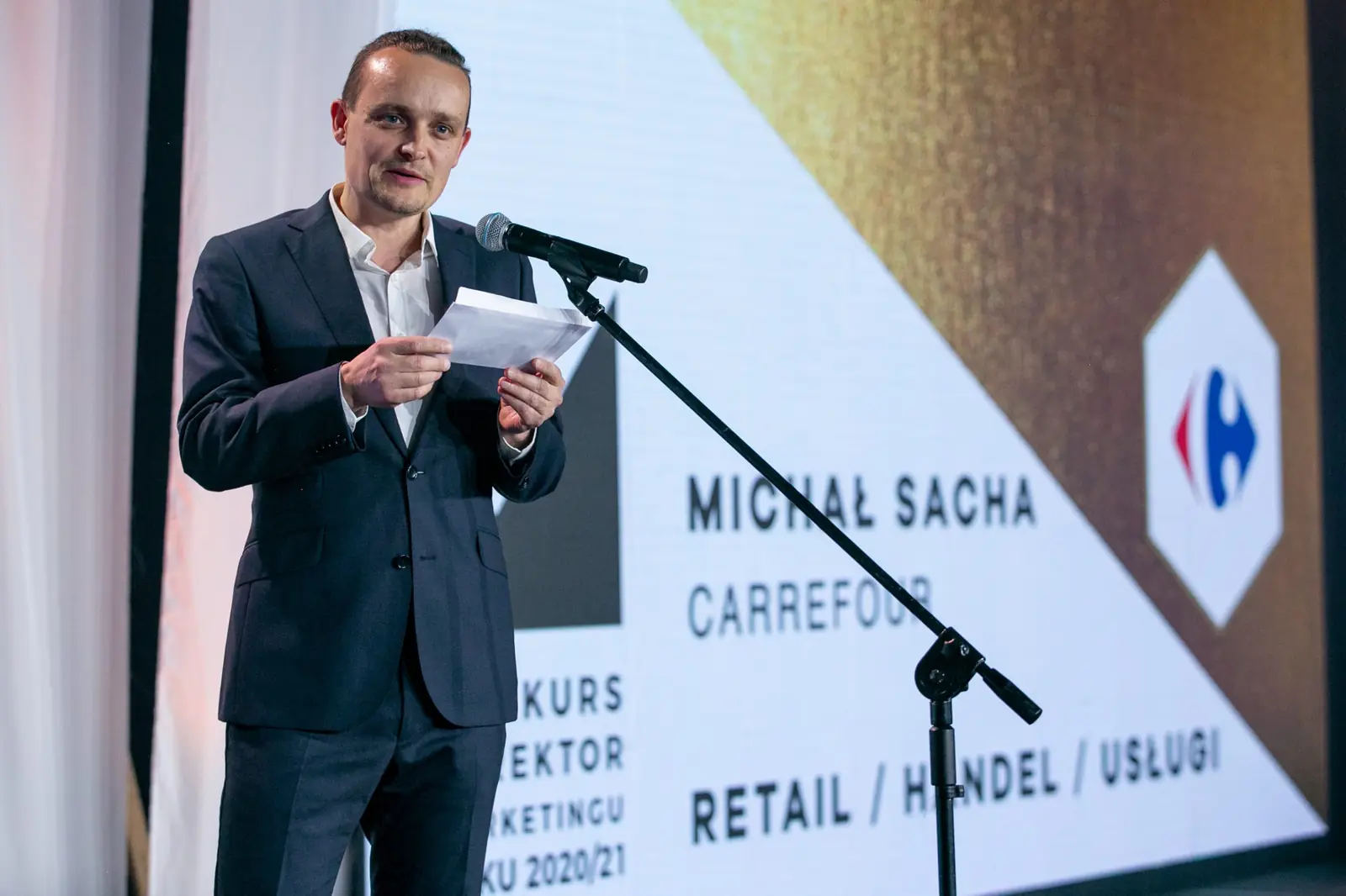 Mężczyzna w ciemnym garniturze czyta z kartki przy mikrofonie w kategorii retail, handel i usługi — Gala DMR X (2021)