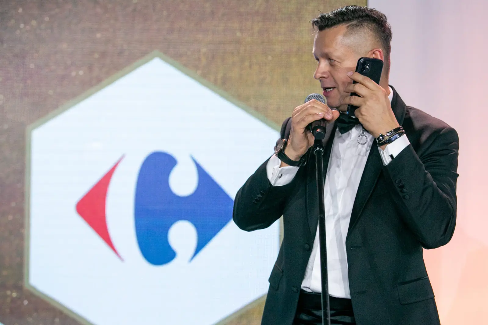 Prowadzący w smokingu rozmawia przez telefon na scenie, trzymając w drugiej dłoni mikrofon — Gala DMR X (2021)