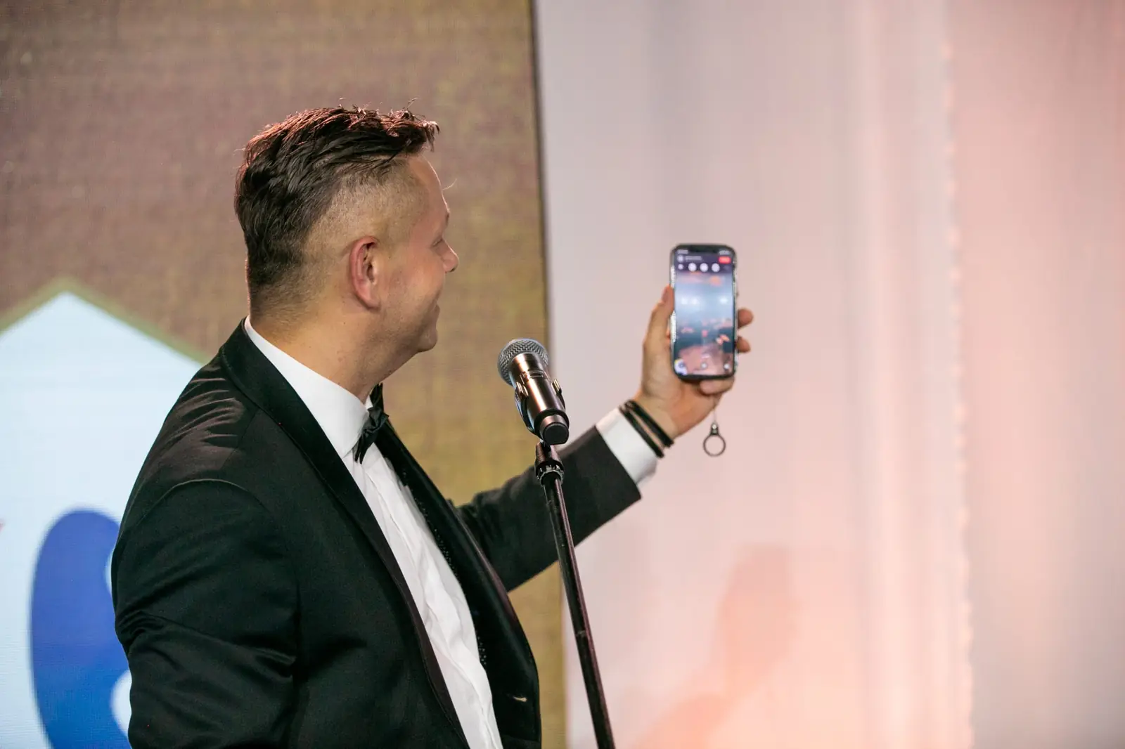 Host holds up his phone with the screen facing the audience while standing at the microphone — DMR Gala X (2021)